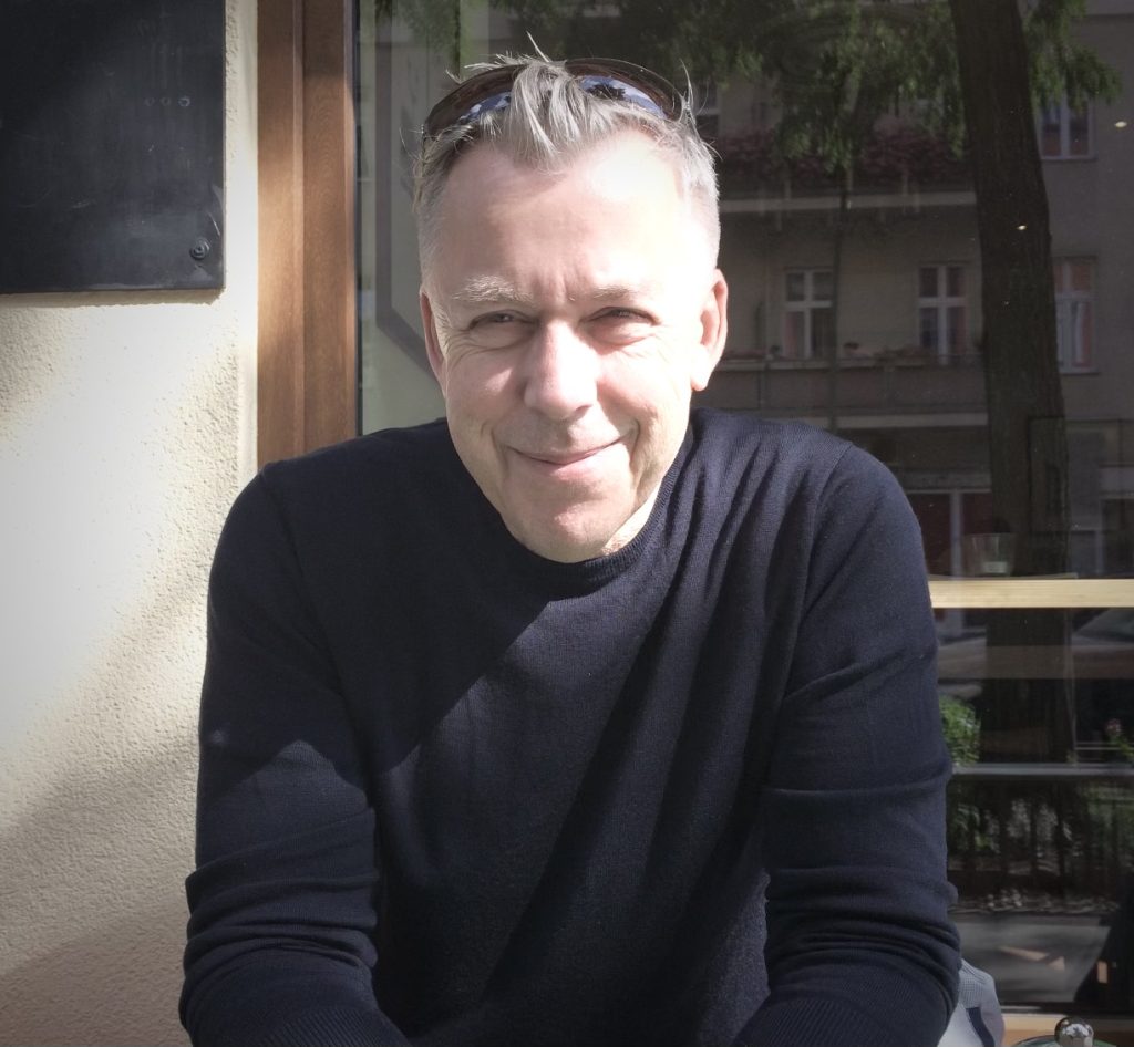 Bild von Christoph Nicolaisen, er sitzt vor einem Café und lächelt in die Kamera.